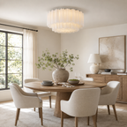 A modern cylindrical chandelier with pleated white glass shades hangs over a round wooden dining table surrounded by six upholstered cream chairs in a bright, contemporary dining room.