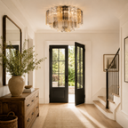 A modern multi-tiered crystal chandelier hangs in a bright, elegant entryway with white walls, black double doors, a wooden console table with a large vase of greenery, and a staircase on the right.