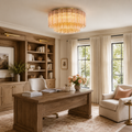 A beautifully styled home office featuring a large wooden writing desk, built-in wooden bookshelves, a cream-colored armchair, and a modern cylindrical crystal chandelier hanging from the ceiling.