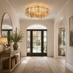 A luxury chandelier with a clear glass cylindrical column design and warm gold-toned lighting, hanging in a bright, elegant entryway with arched doorways, a console table, and a light-colored rug.