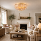 A beautifully styled living room featuring a large, round, illuminated crystal chandelier hanging above a white fireplace mantel. The room is decorated with a cream sofa, a wooden coffee table, a potted olive tree, and soft neutral tones throughout.
