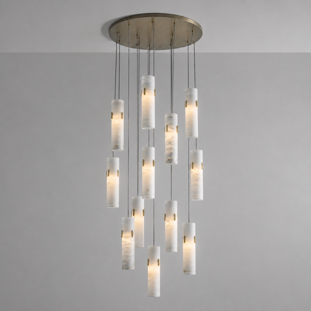 A modern multi-light chandelier pendant fixture with a brushed brass circular ceiling canopy and twelve cylindrical white marble or stone-look glass shades suspended at varying lengths against a plain light gray wall.