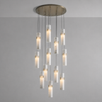 A modern multi-light chandelier pendant fixture with a brushed brass circular ceiling canopy and twelve cylindrical white marble or stone-look glass shades suspended at varying lengths against a plain light gray wall.