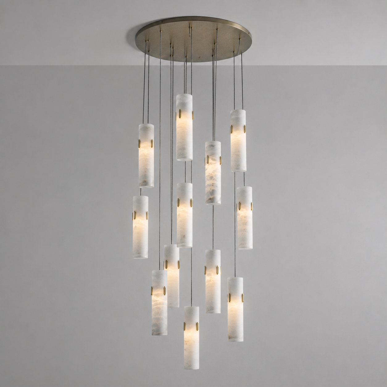 A modern multi-light chandelier pendant fixture with a brushed brass circular ceiling canopy and twelve cylindrical white marble or stone-look glass shades suspended at varying lengths against a plain light gray wall.
