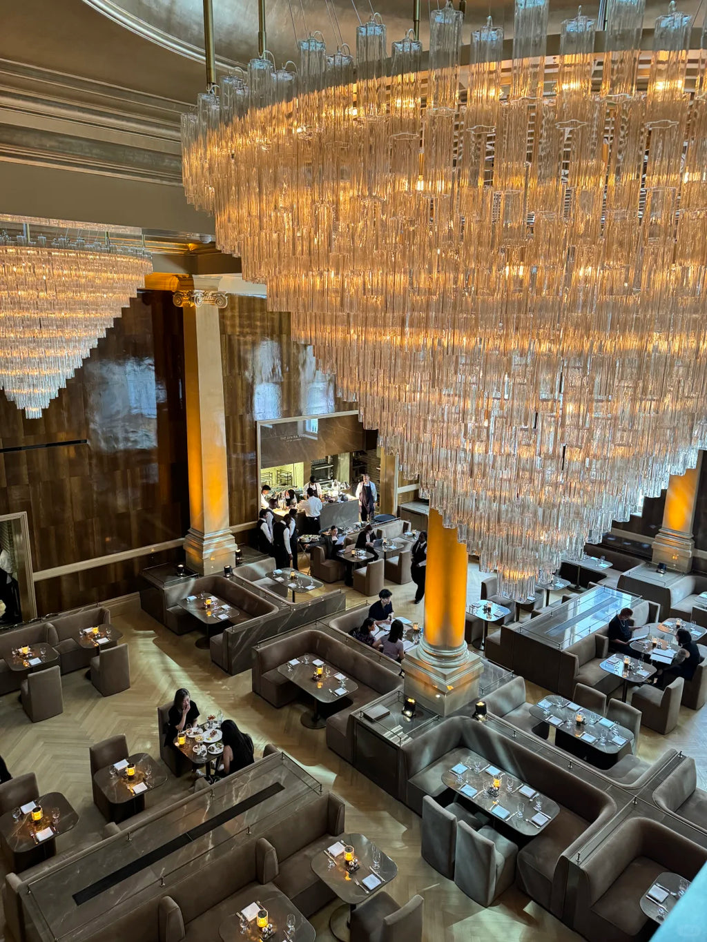 A high-angle view of an elegant, upscale restaurant or lounge interior featuring a massive, glowing crystal chandelier, dark gray upholstered booths, and tables set with candles and glassware.